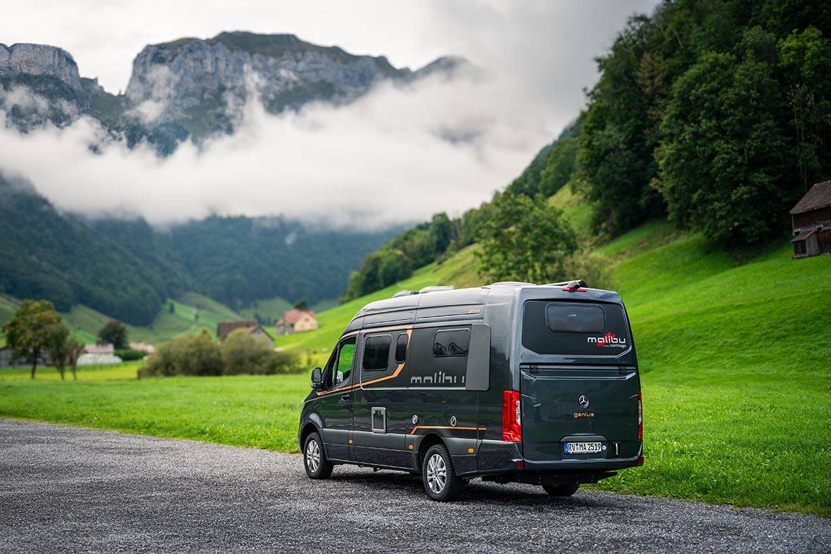 Malibu Genius 4x4 auf nasser Straße in Berglandschaft mit grünen Wiesen und tiefhängenden Wolken.
