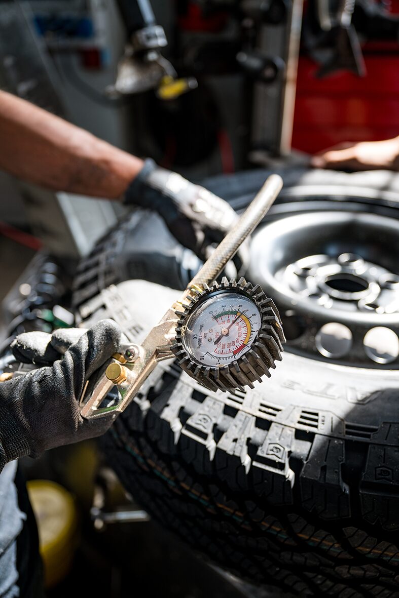 Detailaufnahme eines Mechanikers bei AllRoad, der in einer Werkstatt den Reifendruck eines Geländewagens überprüft. Im Vordergrund ist ein Reifendruckmessgerät zu sehen, während der Reifen eines Campers bearbeitet wird.