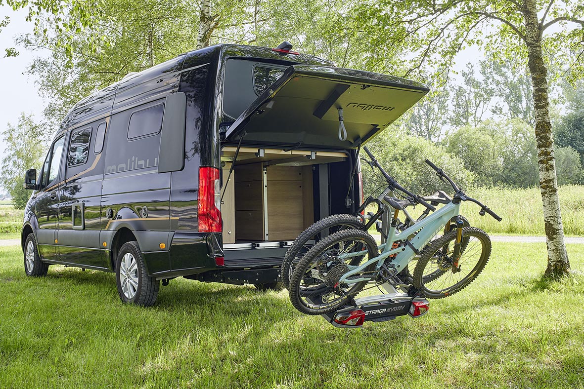 Malibu Genius 4x4 mit geöffnetem Heck, abgeklapptem Fahrradträger und zwei Mountainbikes auf einer Wiese.