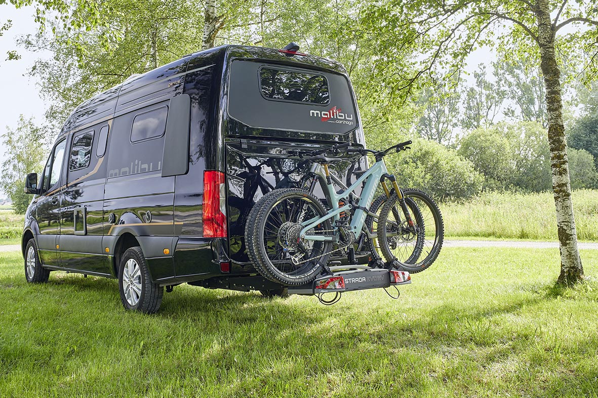 Malibu Genius 4x4 mit Heck-Fahrradträger und zwei Mountainbikes auf einer grünen Wiese.