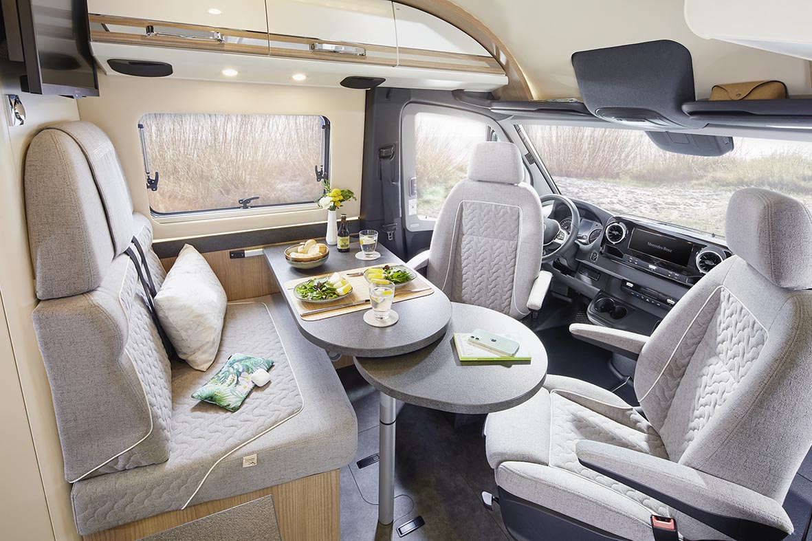 Sitzgruppe im Malibu Genius 4x4 mit drehbaren Fahrerhaussitzen, Tisch und heller Innenausstattung.